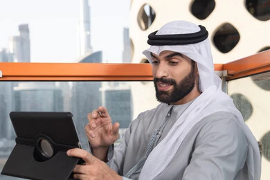 كوفي شوب راقي بالامارات، دمج التقنية والتكنولوجيا في الروتين اليومي، تعابير السعادة و السرور،  صورة مقربه لرجل عربي خليجي اماراتي يرتدي الكندورة و الغتره  يستخدم التابلت في مقهى مرتفع مطل على المدينة