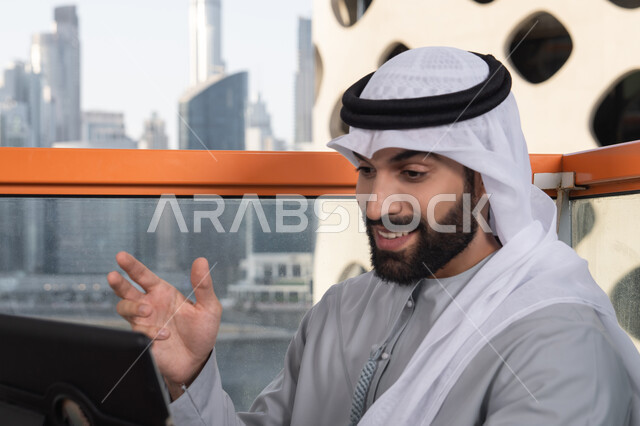 دمج التقنية والتكنولوجيا في الروتين اليومي، تعابير السعادة و السرور،  صورة مقربه لرجل عربي خليجي اماراتي يرتدي الكندورة و الغتره  يستخدم التابلت في مقهى مرتفع مطل على المدينة