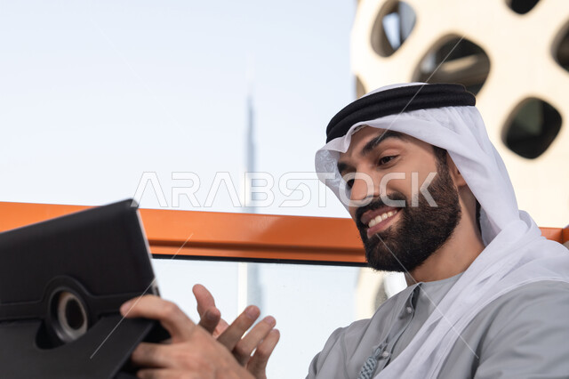تعابير السعادة و السرور، دمج التقنية والتكنولوجيا في الحياة اليومية،  صورة مقربه لرجل عربي خليجي اماراتي يرتدي الكندورة و الغتره  يستخدم التابلت في مقهى مرتفع مطل على المدينة