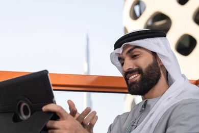Expressions of happiness and joy, integrating technology and tech into daily life, a close-up image of an Emirati Gulf Arab man wearing a kandura and ghutrah using a tablet in a high café overlooking the city.