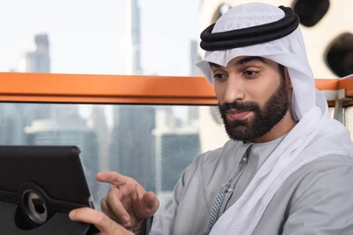 دمج التقنية والتكنولوجيا في الحياة اليومية، تعابير وايماءات الاندماج،  صورة مقربه لرجل عربي خليجي اماراتي يرتدي الكندورة و الغتره  يستخدم التابلت في مقهى مرتفع مطل على المدينة