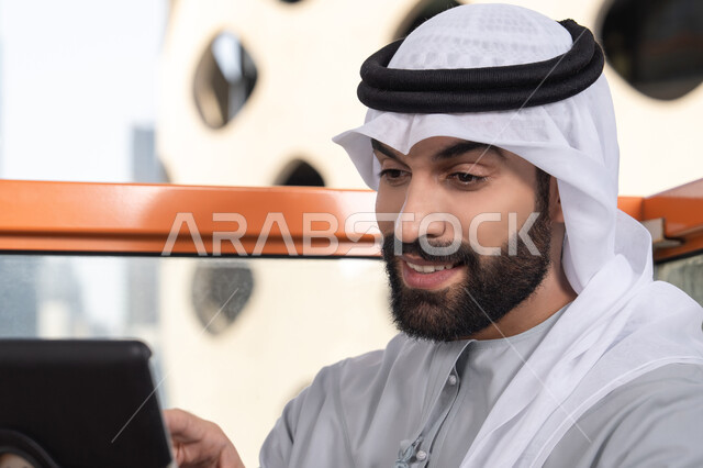 تعابير وايماءات الاندماج،  صورة مقربه لرجل عربي خليجي اماراتي يرتدي الكندورة و الغتره  يستخدم التابلت في مقهى مرتفع مطل على المدينة ، استخدام جهاز تقني حديث ومتطور
