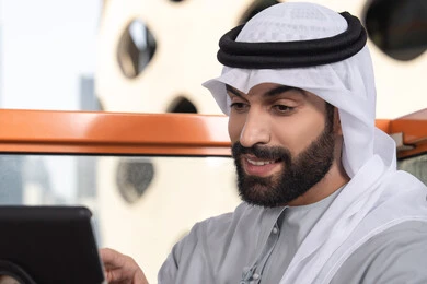 تعابير وايماءات الاندماج،  صورة مقربه لرجل عربي خليجي اماراتي يرتدي الكندورة و الغتره  يستخدم التابلت في مقهى مرتفع مطل على المدينة ، استخدام جهاز تقني حديث ومتطور
