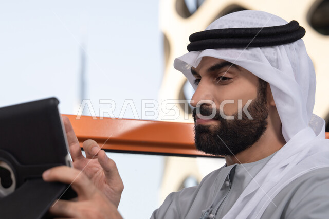 انجاز العمل عن بعد في الهواء الطلق،  صورة مقربه لرجل عربي خليجي اماراتي يرتدي الكندورة و الغتره  يستخدم التابلت في مقهى مرتفع مطل على المدينة ، استخدام جهاز تقني حديث ومتطور