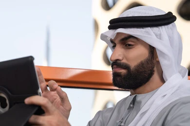 Accomplishing remote work outdoors, a close-up image of an Emirati Gulf Arab man wearing a kandura and ghutrah using a tablet in a high café overlooking the city, utilizing a modern and advanced technological device.