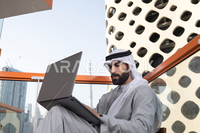 Working to achieve goals and strategies, completing remote work tasks with integration gestures, managing and organizing business affairs, using modern and advanced technology, an Arab Gulf Emirati man wearing a kandura and a white ghutrah using a laptop.