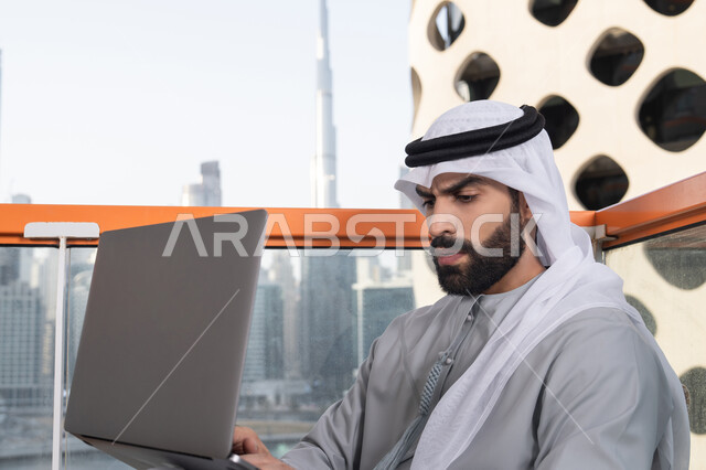 دمج العمل مع التقنية والتكنولوجيا الحديثة، انجاز مهام العمل عن بعد بايماءات الاندماج، إدارة شؤون الأعمال وتنظيمها ، استخدام جهاز تقني حديث ومتطور، رجل عربي خليجي اماراتي يرتدي الكندورة والغترة البيضاء يستخدم الحاسوب المحمول
