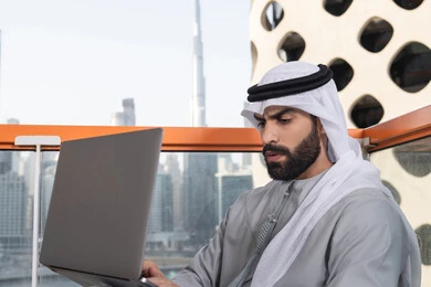دمج العمل مع التقنية والتكنولوجيا الحديثة، انجاز مهام العمل عن بعد بايماءات الاندماج، إدارة شؤون الأعمال وتنظيمها ، استخدام جهاز تقني حديث ومتطور، رجل عربي خليجي اماراتي يرتدي الكندورة والغترة البيضاء يستخدم الحاسوب المحمول