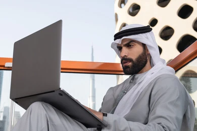 Completing remote work tasks with integration gestures, communicating with family and friends via laptop, managing and organizing business affairs, using a modern and advanced technical device, an Arab Gulf Emirati man wearing a kandura and a white ghutrah using a laptop.