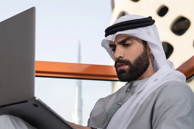 التواصل مع الأهل والاصدقاء عبر اللابتوب ، إنجاز مهام العمل عن بعد، إدارة شؤون الأعمال وتنظيمها ، استخدام جهاز تقني حديث ومتطور، رجل عربي خليجي اماراتي يرتدي الكندورة والغترة البيضاء يستخدم الحاسوب المحمول