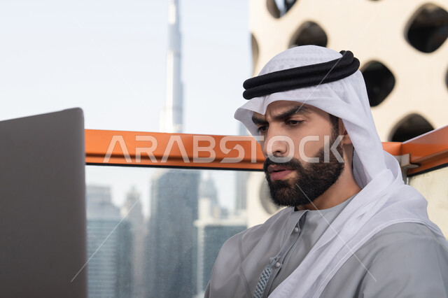 Gestures of engagement and focus while working, accomplishing remote work tasks, managing and organizing business affairs via laptop, using a modern and advanced technical device, an Emirati Gulf Arab man wearing a kandura and a white ghutrah using a laptop.