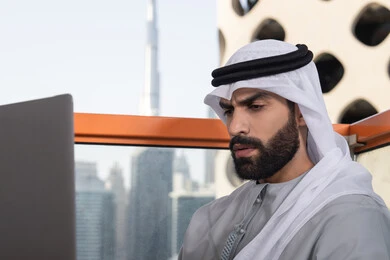 إيماءات الاندماج والتركيز أثناء العمل ، إنجاز مهام العمل عن بعد، إدارة شؤون الأعمال وتنظيمها عبر اللابتوب، استخدام جهاز تقني حديث ومتطور، رجل عربي خليجي اماراتي يرتدي الكندورة والغترة البيضاء يستخدم الحاسوب المحمول
