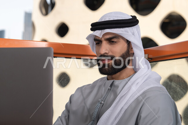 Gestures of happiness and joy, accomplishing remote work tasks, managing and organizing business affairs via laptop, using a modern and advanced technical device, an Arab Gulf Emirati man wearing a kandura and a white ghutrah using a laptop.