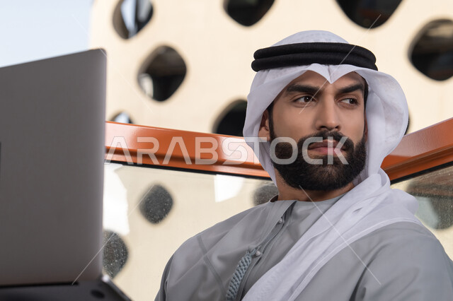 إدارة شؤون الأعمال وتنظيمها عبر اللابتوب، استخدام جهاز تقني حديث ومتطور، إنجاز مهام العمل عن بعد، رجل عربي خليجي اماراتي يرتدي الكندورة والغترة البيضاء يستخدم الحاسوب المحمول و ينظر الى شيء ما