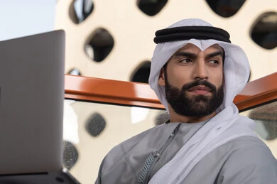 إدارة شؤون الأعمال وتنظيمها عبر اللابتوب، استخدام جهاز تقني حديث ومتطور، إنجاز مهام العمل عن بعد، رجل عربي خليجي اماراتي يرتدي الكندورة والغترة البيضاء يستخدم الحاسوب المحمول و ينظر الى شيء ما
