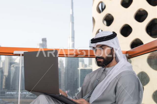 استخدام جهاز تقني حديث ومتطور، إنجاز مهام العمل عن بعد، إدارة شؤون الأعمال وتنظيمها عبر اللابتوب، ايماءات التركيز و الاندماج، رجل عربي خليجي اماراتي يرتدي الكندورة والغترة البيضاء يستخدم الحاسوب المحمول