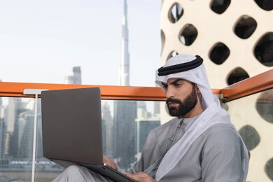 استخدام جهاز تقني حديث ومتطور، إنجاز مهام العمل عن بعد، إدارة شؤون الأعمال وتنظيمها عبر اللابتوب، ايماءات التركيز و الاندماج، رجل عربي خليجي اماراتي يرتدي الكندورة والغترة البيضاء يستخدم الحاسوب المحمول