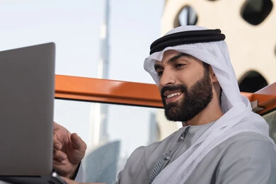 إنجاز مهام العمل عن بعد، إدارة شؤون الأعمال وتنظيمها عبر اللابتوب، ايماءات السعادة والسرور، استخدام جهاز تقني حديث ومتطور، رجل عربي خليجي اماراتي يرتدي الكندورة والغترة البيضاء يستخدم الحاسوب المحمول