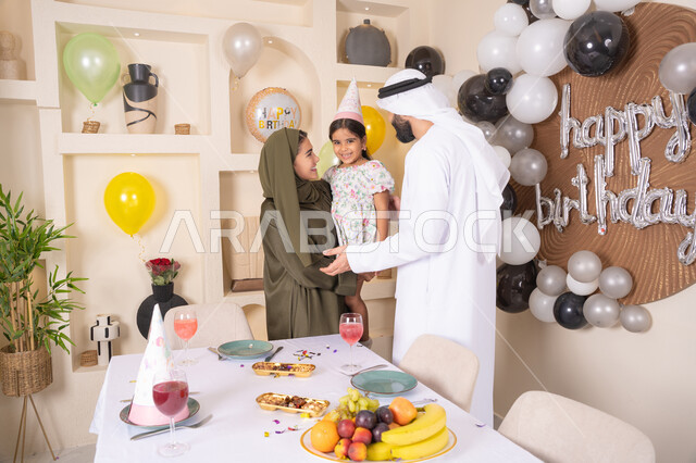 مفهوم الحب والالفه والموده، عائله عربيه خليجيه اماراتية تحتفل بعيد ميلاد ابنتهم ، ام اماراتيه تحمل ابنتها لاعلى بتعابير البهجة، مشاركة لأجواء رائعة عائلية داخل المنزل، تعليق الزينة والبالونات الملونة ، ايماءات السعاده والسرور ، اعياد ومناسبات سعيدة،  مشاعر الحب والحنان في اجتماع عائلي سعيد