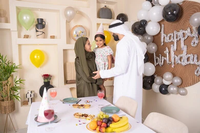 The concept of love, intimacy and affection, an Arab Gulf Emirati family celebrating their daughter's birthday, an Emirati mother carrying her daughter up with expressions of joy, sharing a wonderful family atmosphere inside the house, hanging colorful decorations and balloons, gestures of happiness and pleasure, happy holidays and occasions, feelings of love and tenderness in a happy family gathering
