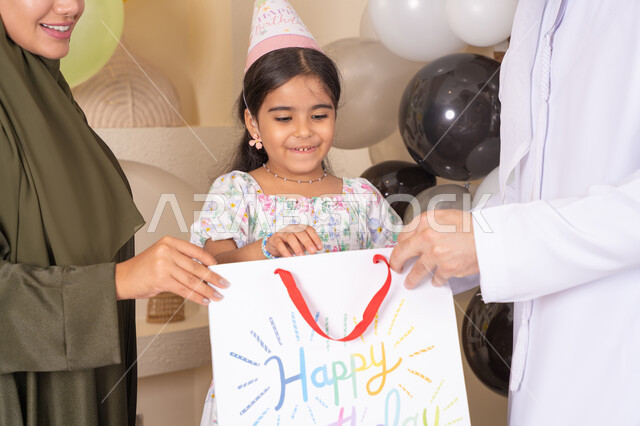 Hanging colorful decorations and balloons, expressions of surprise, happiness, and joy, sharing a wonderful family atmosphere at home, a close-up photo of an Emirati Gulf Arab family celebrating their daughter's birthday and presenting her with a gift, happy holidays and occasions, giving gifts and surprises, the concept of love, intimacy, and affection.