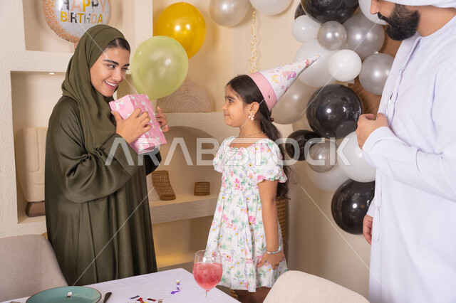 ام اماراتيه تقوم بتقديم الهديه لابنتها ، تعابير المفاجأه والسعادة و السرور، عائله عربيه خليجيه اماراتية تحتفل بعيد ميلاد ابنتهم ،اعياد ومناسبات سعيدة، تقديم الهدايا والمفاجآت للأطفال، مفهوم الحب والالفه والموده،مشاركة لأجواء رائعة عائلية داخل المنزل، تعليق الزينة والبالونات الملونة