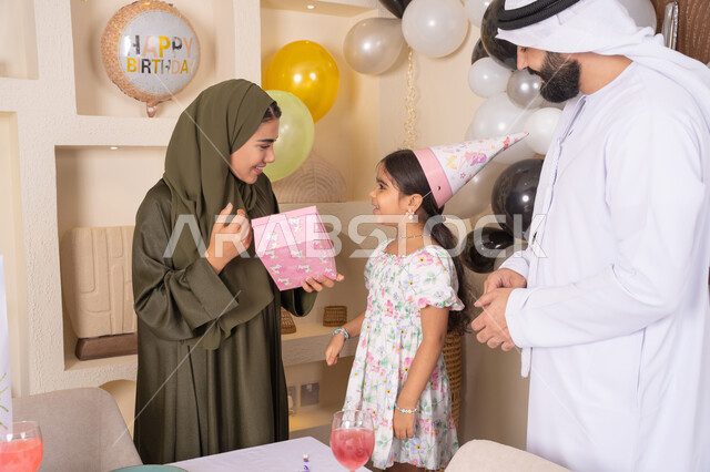 Gestures of happiness and joy, sharing a wonderful family atmosphere inside the home, an Arab Gulf Emirati family celebrating their daughter's birthday, happy holidays and occasions, hanging decorations and colorful balloons, an Emirati mother presenting a gift to her daughter, the concept of love, intimacy and affection, presenting gifts and surprises to children