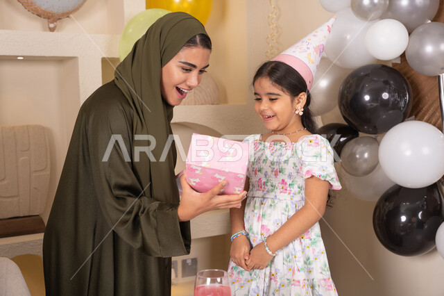 Hanging colorful decorations and balloons, an Emirati mother presenting a gift to her daughter, the concept of love, intimacy and affection, presenting gifts and surprises to children, happy holidays and occasions, gestures of happiness and pleasure, sharing a wonderful family atmosphere inside the home, an Arab Gulf Emirati family celebrating their daughter's birthday