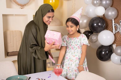 Emirati mother presents a gift to her daughter, the concept of love, intimacy and affection, presenting gifts and surprises to children, happy holidays and occasions, hanging decorations and colorful balloons, gestures of happiness and pleasure, sharing a wonderful family atmosphere inside the home, an Arab Gulf Emirati family celebrating their daughter's birthday