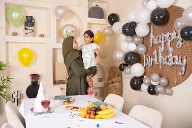 ام عربية خليجية تحمل ابنتها  للاعلى بايماءات السعادة و السرور، عائله عربيه خليجيه اماراتية تقوم بالاحتفال بعيد ميلاد ابنتهم ، تقديم الهدايا والمفاجآت للأطفال، تعليق الزينة والبالونات الملونة، اعياد ومناسبات سعيدة
