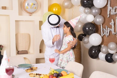 Giving gifts and surprises to children, happy holidays and occasions, hanging decorations and colorful balloons, an Arab Gulf father embracing his daughter with gestures of happiness and pleasure, an Arab Gulf Emirati family celebrating their daughter's birthday