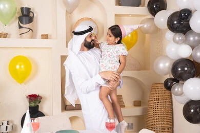 اعياد ومناسبات سعيدة، تعليق الزينة والبالونات الملونة،  تقديم الهدايا والمفاجآت للأطفال،اب عربي خليجي يقوم بحمل طفلته للاعلي بايماءات السعادة و السرور، عائله عربيه خليجيه اماراتية تقوم بالاحتفال بعيد ميلاد ابنتهم