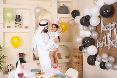 تعليق الزينة والبالونات الملونة،  تقديم الهدايا والمفاجآت للأطفال، اعياد ومناسبات سعيدة، اب عربي خليجي يقوم بحمل طفلته للاعلي بايماءات السعادة و السرور، عائله عربيه خليجيه اماراتية تقوم بالاحتفال بعيد ميلاد ابنتهم