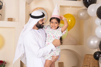 تقديم الهدايا والمفاجآت للأطفال، تعليق الزينة والبالونات الملونة، اعياد ومناسبات سعيدة، اب عربي خليجي يقوم بحمل طفلته للاعلي بايماءات السعادة و السرور، عائله عربيه خليجيه اماراتية تقوم بالاحتفال بعيد ميلاد ابنتهم