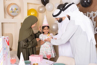 Feelings of love and tenderness in a happy family gathering, balloons and a birthday cake decorated with candles, an Emirati Gulf Arab family celebrating their daughter's birthday, gestures of joy and happiness, sharing and giving gifts on occasions and birthdays, a family atmosphere full of intimacy