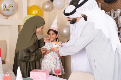 بلالين وكعكة يوم الميلاد المزينة بالشموع ، عائلة عربية خليجية إماراتية تحتفل بعيد ميلاد ابنتهم ، إيماءات الفرح و السعادة، مشاعر الحب والحنان في اجتماع عائلي سعيد، مشاركة وتقديم الهدايا بالمناسبات وأعياد الميلاد، أجواء أسرية مليئة بالألفة
