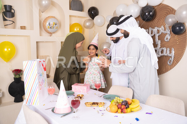 مشاركة وتقديم الهدايا بالمناسبات وأعياد الميلاد، مشاعر الحب والحنان في اجتماع عائلي سعيد، إيماءات الفرح و السعادة، عائلة عربية خليجية إماراتية تحتفل بعيد ميلاد ابنتهم ،أجواء أسرية مليئة بالألفة، بلالين وكعكة يوم الميلاد المزينة بالشموع
