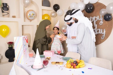 مشاركة وتقديم الهدايا بالمناسبات وأعياد الميلاد، مشاعر الحب والحنان في اجتماع عائلي سعيد، إيماءات الفرح و السعادة، عائلة عربية خليجية إماراتية تحتفل بعيد ميلاد ابنتهم ،أجواء أسرية مليئة بالألفة، بلالين وكعكة يوم الميلاد المزينة بالشموع