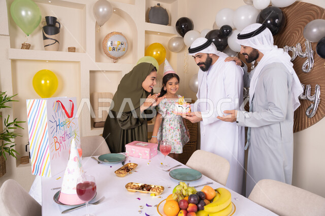 An Emirati Gulf Arab family having a good time at home, gestures of joy and happiness, feelings of love and tenderness in a happy family gathering, sharing and offering gifts on occasions and birthdays, a family atmosphere full of intimacy, balloons and a birthday cake decorated with candles