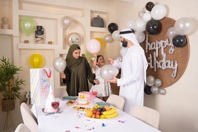 مشاركة وتقديم الهدايا بالمناسبات وأعياد الميلاد، إيماءات الفرح و السعادة، مشاعر الحب والحنان في اجتماع عائلي سعيد, أجواء أسرية مليئة بالألفة، بلالين وكعكة يوم الميلاد المزينة بالشموع،عائلة عربية خليجية إماراتية تقضي أوقات ممتعه في المنزل وتلعب بالبلالين
