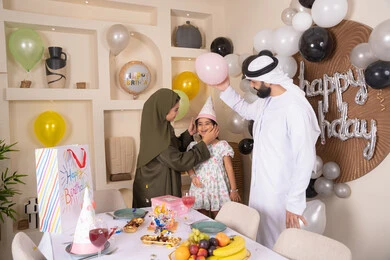 Balloons and a birthday cake decorated with candles, a family atmosphere full of intimacy, feelings of love and tenderness in a happy family gathering, an Emirati Gulf Arab family spending fun times at home and playing with balloons, gestures of joy and happiness, sharing and giving gifts on occasions and birthdays