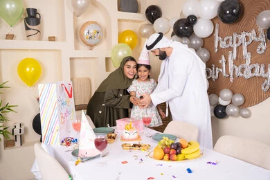 Balloons and a birthday cake decorated with candles, a family atmosphere full of intimacy, feelings of love and tenderness in a happy family gathering, an Arab Gulf Emirati family spending enjoyable times at home, gestures of joy and happiness, sharing and offering gifts on occasions and birthdays