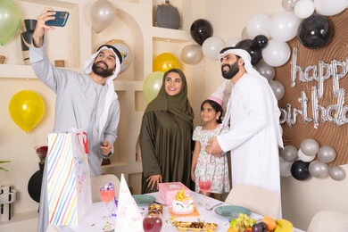 رجل عربي خليجي اماراتي يقوم بأخذ صورة تذكارية من خلال الجوال مع الكعكة المزينة بالشموع، مشاعر الحب والحنان في اجتماع عائلي سعيد, النظر الى الكاميرا بايماءات السرور والفرح، الاحتفال بالمناسبات والأعياد بأجواء عائلية سعيدة, أسرة عربية خليجية اماراتية تقوم بالاحتفال بعيد الميلاد