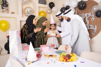 A family atmosphere filled with intimacy, feelings of love and tenderness in a happy family gathering, balloons and a birthday cake decorated with candles, an Arab Gulf Emirati family having a good time at home, gestures of joy and happiness, sharing and offering gifts on occasions and birthdays.