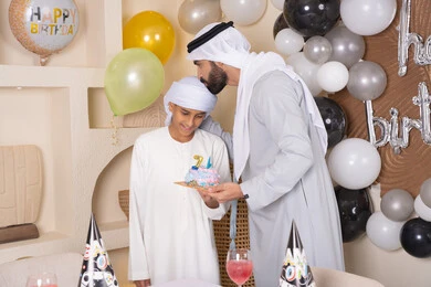 An Emirati Gulf Arab father celebrating and kissing his son's head. Birthday decorations, gestures of happiness and joy, the concept of dedication and appreciation, happy family holidays and occasions, an Emirati Gulf Arab family having a good time.