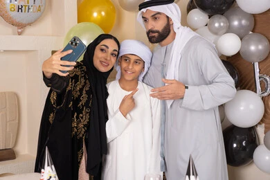Feelings of love and tenderness in a happy family gathering, an Arab Gulf Emirati family celebrating a birthday, an Arab Gulf Emirati mother taking a souvenir photo with her mobile phone with a cake decorated with candles, celebrating occasions and holidays in a happy family atmosphere, looking at the camera with gestures of pleasure and joy