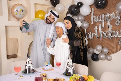 An Emirati Gulf Arab man takes a souvenir photo with his mobile phone with a cake decorated with candles, celebrating occasions and holidays in a happy family atmosphere, looking at the camera with gestures of pleasure and joy, feelings of love and tenderness in a happy family gathering, an Emirati Gulf Arab family celebrating a birthday