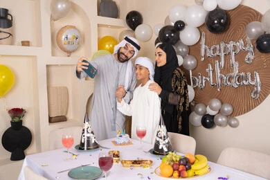 النظر الى الكاميرا بايماءات السرور والفرح ، مشاعر الحب والحنان في اجتماع عائلي سعيد,  أسرة عربية خليجية اماراتية تقوم بالاحتفال بعيد الميلاد، الاحتفال بالمناسبات والأعياد بأجواء عائلية سعيدة, رجل عربي خليجي اماراتي يقوم بأخذ صورة تذكارية من خلال الجوال مع الكعكة المزينة بالشموع