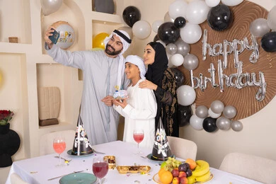 أسرة عربية خليجية اماراتية تقوم بالاحتفال بعيد الميلاد، الاحتفال بالمناسبات والأعياد بأجواء عائلية سعيدة, رجل عربي خليجي اماراتي يقوم بأخذ صورة تذكارية من خلال الجوال مع الكعكة المزينة بالشموع، مشاعر الحب والحنان في اجتماع عائلي سعيد, النظر الى الكاميرا بايماءات السرور والفرح