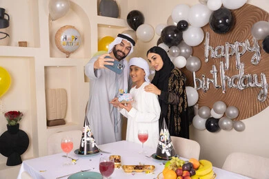 الاحتفال بالمناسبات والأعياد بأجواء عائلية سعيدة, أسرة عربية خليجية اماراتية تقوم بالاحتفال بعيد الميلاد، رجل عربي خليجي اماراتي يقوم بأخذ صورة تذكارية من خلال الجوال مع الكعكة المزينة بالشموع، مشاعر الحب والحنان في اجتماع عائلي سعيد, النظر الى الكاميرا بايماءات السرور والفرح
